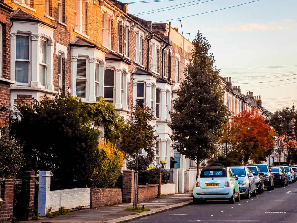 Cars parked outside orange brick houses on a tree-lined W12 street in autumn