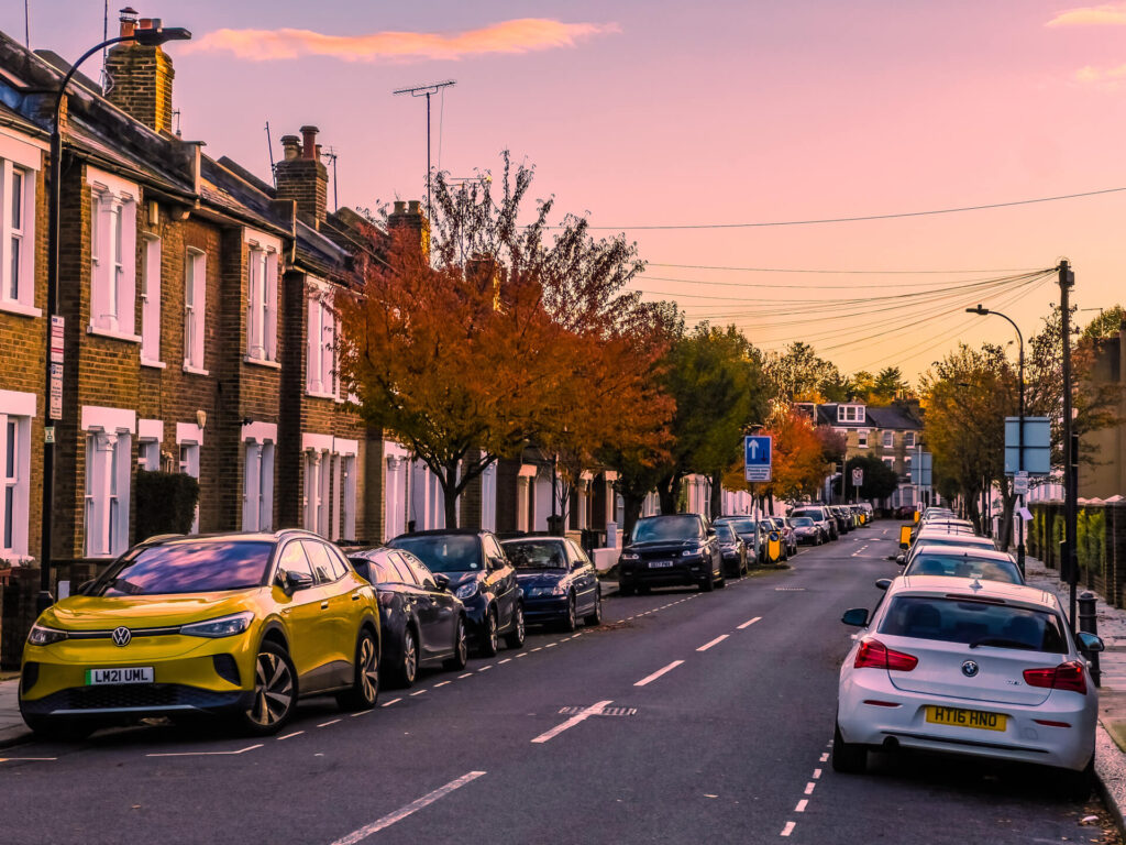 Cars parked outside orange brick houses on a tree-lined W12 street in autumn