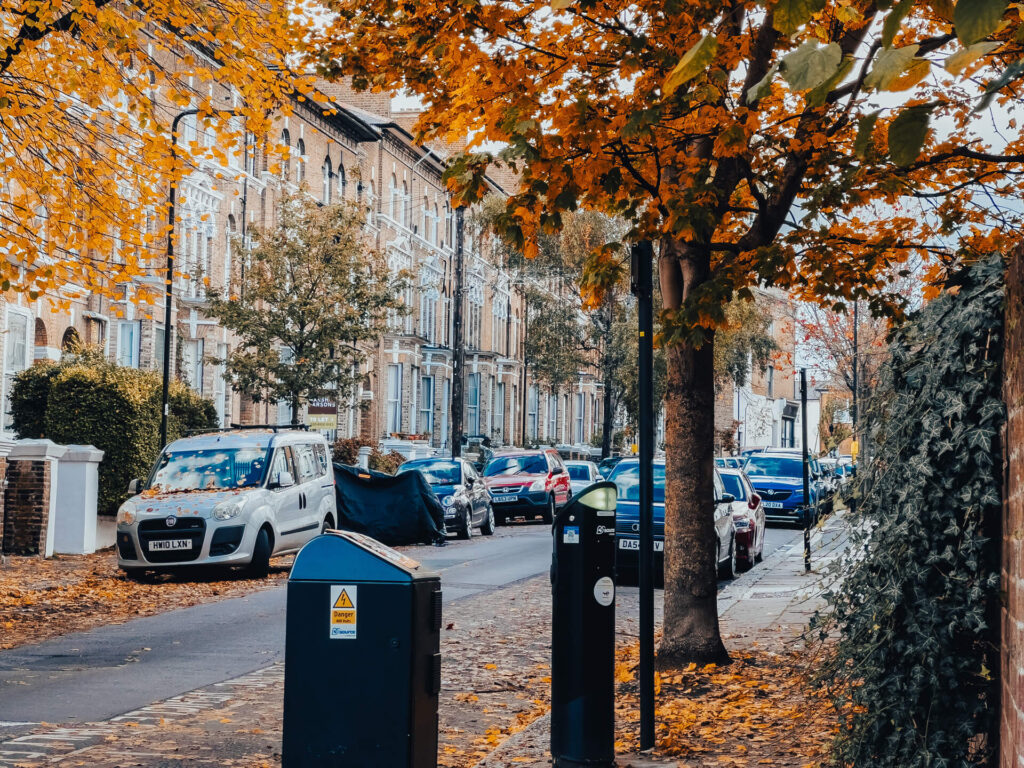 Cars parked outside orange brick houses on a tree-lined W12 street in autumn