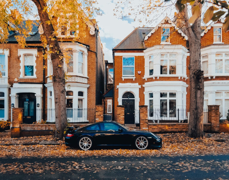 A black Porsche parked on top of a bed of autumn leaves set in front of a street of orange brick houses in W4